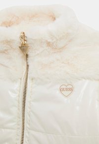Giacca puffer bianca con un morbido colletto in pelliccia sintetica. Presenta una chiusura con zip, dettagli in oro e una toppa a forma di cuore sul petto.