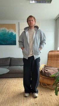 Sudadera gris con cremallera y forro blanco, camiseta ligera, pantalones deportivos oscuros y zapatos grises. Persona de pie con las manos en los bolsillos, en un entorno interior.