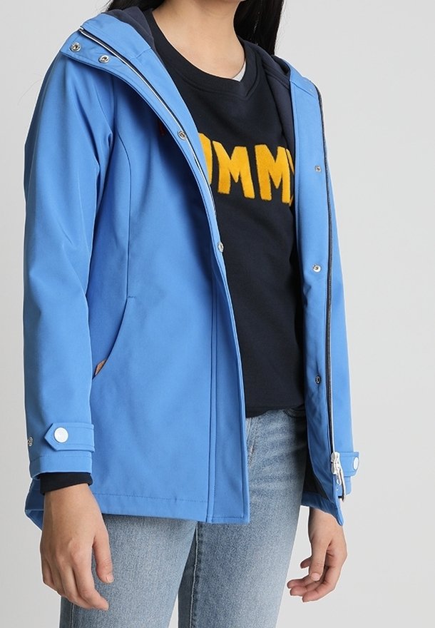 Persona con una chaqueta azul clara sobre una sudadera negra con letras amarillas, combinada con jeans azul claro, de pie contra un fondo plano.