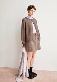 Brauner Strickcardigan mit gerippter Textur, über einem helllila Rollkragenpullover und einem braunen Tweedrock getragen, kombiniert mit schwarzen, fransigen Schuhen.