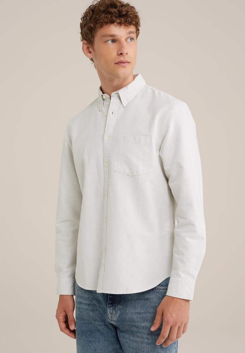 Chemise boutonnée à manches longues de couleur claire avec une poche avant, tissu texturé et ourlet arrondi, associée à un jean bleu.