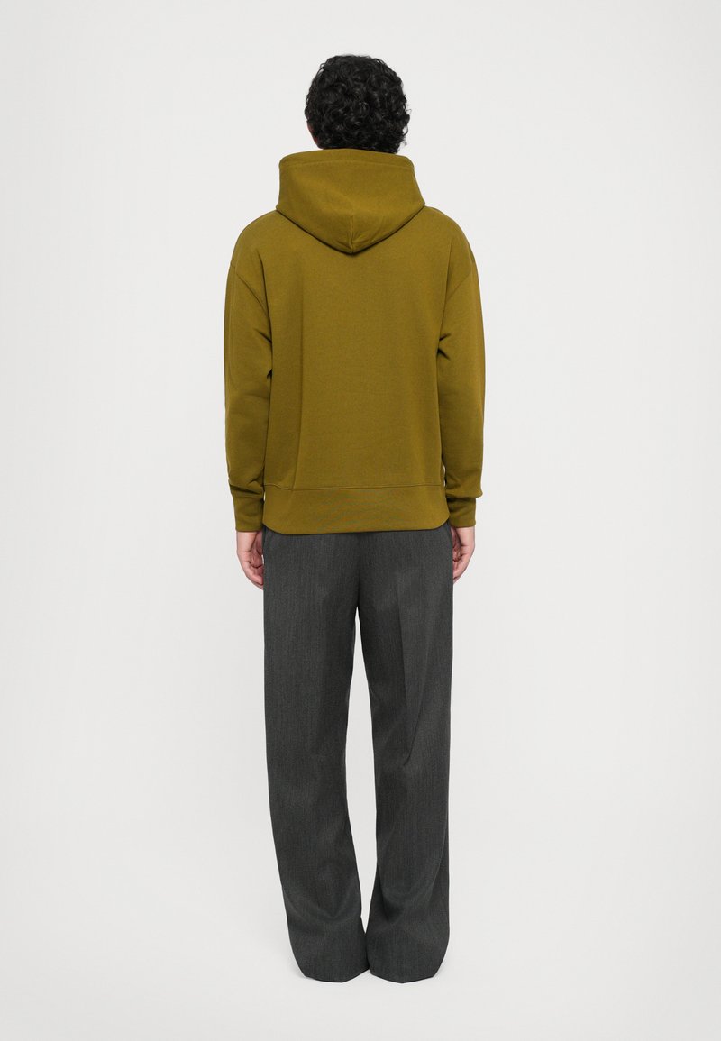 Personne portant un sweat à capuche vert olive et un pantalon gris foncé ample, se tenant de dos face à la caméra contre un fond blanc uni.
