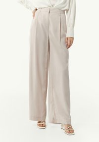 Jasnoróżowe spodnie w stylu wide-leg, wykonane z miękkiego materiału, z wysokim stanem, plisami i luźnym krojem, w zestawie z białą koszulą z długim rękawem.