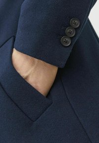 Mörkblå kavaj i ullblandning med en skarp krage, en sidoficka och tre svarta knappar på ärmen. Texturerad tyg med en slät yta.
