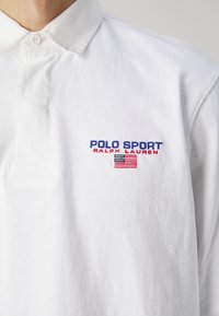Vit bomullspolo med knäppt krage och broderad "POLO SPORT" samt flaggadetalj i blått och rött på vänster bröst.