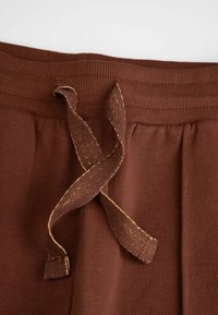 Brązowe bawełniane spodnie dresowe z prążkowanym pasem i złotymi akcentowanymi sznurkami. Posiadają subtelną teksturę i boczne plisy dla komfortu.