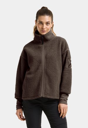 Jeune femme à la peau claire et aux cheveux tirés en arrière portant une veste polaire marron foncé zippée et un pantalon noir sur un fond uni.