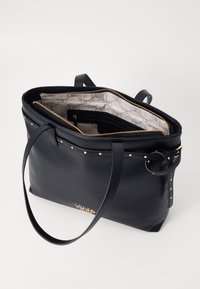 Bolso tote de cuero negro con herrajes dorados, cierre superior con cremallera, acentos de tachuelas, bolsillos interiores y un forro estampado. Diseño espacioso y estructurado.