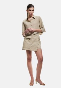 Khaki knap-up skjorte med oprullede ærmer, parret med en højtskåret omslagsskjørt med en lomme. Model iført open-toe hæle sandaler.