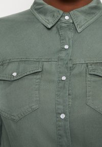 Vaaleanvihreä napitettava paita, joka on valmistettu pehmeästä kankaasta. Paidassa on kaulus, kaksi rintataskua läpällä ja hopeiset nepparit.