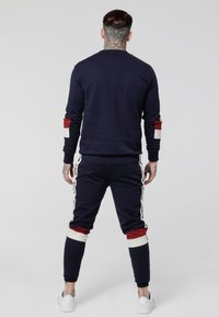 Sada navy modré mikiny a tepláků, s červenými a bílými akcenty na rukávech a kalhotách, s logo pruhy na stranách. Bavlněný materiál.