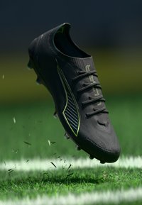 Crampons de football noirs avec un dessus texturé, accents vert fluo, six œillets et crampons moulés, flottant au-dessus d'herbe avec de petits fragments de brins.