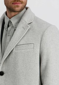 Lichtgrijze wollen blend jas met een geknipte kraag, enkele knoopsluiting, voorkantzak met stikseldetails, gelaagd over een groene button-up shirt.