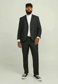 Fato cinza escuro com um casaco de um só botão, golas notched e calças combinando. Camisa branca com botões. Ténis pretos. Acabamento de tecido suave.