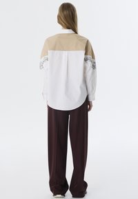 Camisa bloqueada en blanco y beige con intrincados bordados florales negros en las mangas. Corte relajado; dobladillo curvado; combinada con pantalones amplios burdeos.