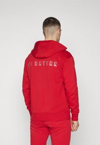 Felpa rossa realizzata in tessuto morbido con tasca a marsupio, caratterizzata da "FC BAYERN" in lettere bianche sul retro e cappuccio con coulisse.