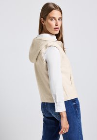 Gilet en polaire beige avec une capuche, sans manches, poches latérales et surface texturée, porté sur une chemise blanche à manches longues et un jean bleu.