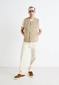 Béžová prošívaná vesta s patentkami, nošená přes bílý žebrovaný tričko. V kombinaci se krémovými kalhotami a hnědými šněrovacími botami. Nastavitelná kšiltovka.