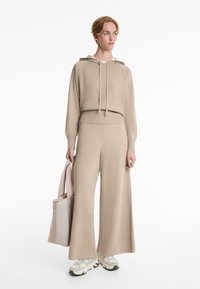 Beige knitted hoodie with ribbed detailing and drawstrings, paired with wide-leg trousers. Slim tote bag and chunky sneakers complete the look.