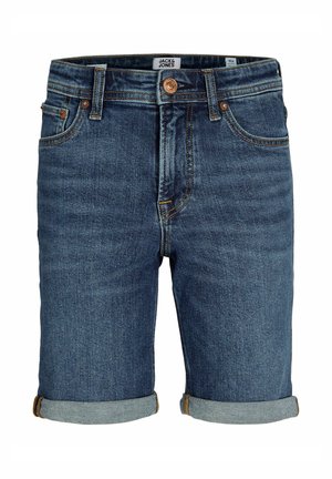 Fiú áll, tengerészkék pólóingben, felhajtott farmer rövidnadrágban, fehér zokniban és fehér tornacipőben, világos egyszínű háttér előtt.