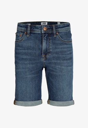 Fiú áll, tengerészkék pólóingben, felhajtott farmer rövidnadrágban, fehér zokniban és fehér tornacipőben, világos egyszínű háttér előtt.