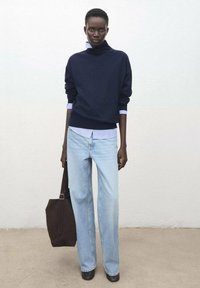Granatowy dzianinowy golf nałożony na niebiesko-białą koszulę w paski, zestawiony z jasnoniebieskimi szerokimi jeansami i ciemnobrązową torbą na ramię.
