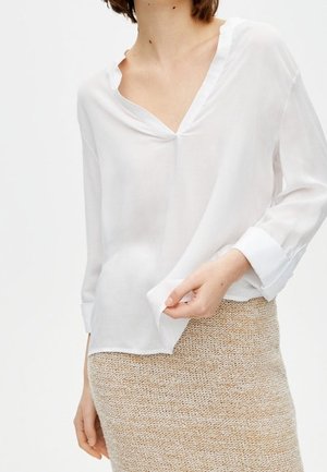 Kobieta ubrana w półprzezroczystą białą bluzkę z dekoltem w serek i podwiniętymi mankietami, zestawioną z beżową spódnicą dzianinową o fakturze.