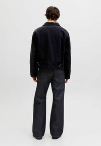 Person med kort krøllet hår iført en sort jakke og mørke, vide jeans, stående med ryggen mod en ensfarvet lys baggrund.