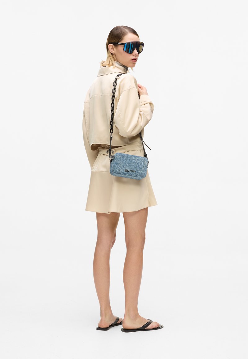 Γυναίκα που φοράει μπεζ cropped σακάκι και φούστα, μαύρα σανδάλια, τεράστια μπλε γυαλιά ηλίου, κρατώντας μικρή μπλε τσάντα με μαύρο λουρί αλυσίδας.