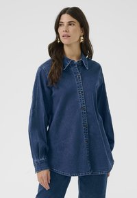 Mujer con cabello largo castaño, vistiendo una camisa de mezclilla oscura abotonada y pantalones a juego, mirando ligeramente de lado frente a un fondo liso.