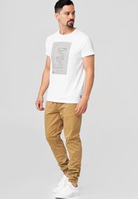 Camiseta blanca con un diseño gráfico lineal en negro, combinada con pantalones jogger color beige, con puños elásticos y zapatillas blancas.