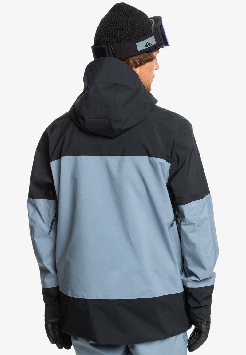 Wodoodporna kurtka z czarno-niebieskim blokowym wzorem. Posiada kaptur, kieszenie na zamek i teksturowany materiał.