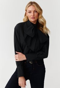 Blusa nera con un ampio fiocco al colletto, maniche lunghe e una superficie liscia, abbinata a jeans scuri. Design semplice ed elegante.