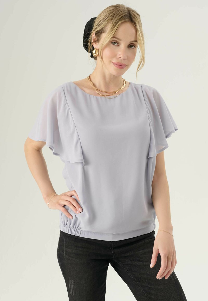Donna con capelli biondi in una fascia nera che indossa orecchini a cerchio dorati, collane a strati, una blusa grigia chiara con maniche a sbuffo e jeans neri.