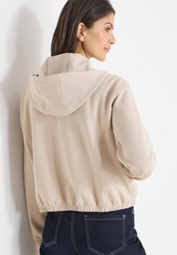 Veste beige à capuche avec ourlet élastique, tissu lisse et détails de coutures minimales. Portée avec un pantalon foncé, vue de dos.