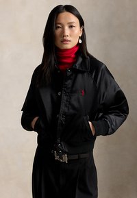 Frau mit langen dunklen Haaren trägt schwarze Satinjacke über rotem Rollkragenpullover, Hände in den Taschen, steht vor neutralem beigefarbenem Hintergrund.