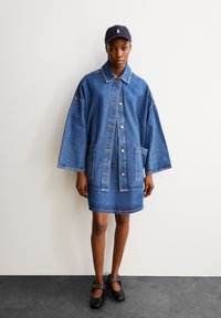 Denim jacket with wide sleeves, dark blue fabric, two front pockets, and button closure, paired with a matching denim skirt and flat shoes.