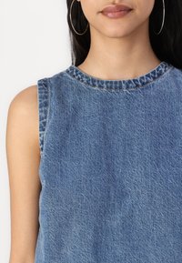 Mujer vestida con una blusa sin mangas de denim azul y grandes pendientes de aro, con brillo labial neutro y cabello largo y oscuro.