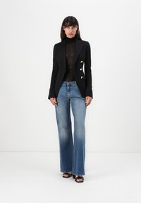 Zwarte dubbelgeknoopte blazer met gouden knopen, doorschijnende zwarte coltrui en lichtblauwe flared jeans. G gedragen met zwarte hakken.