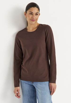 Frau mit dunklem zurückgebundenem Haar, die ein braunes Langarmshirt und hellblaue Jeans trägt, lehnt an einer weißen Wand.