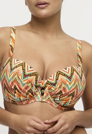 Vrouw draagt een bh met een meerkleurig zigzagpatroon en kralen in het midden, handen losjes in elkaar gevouwen ter hoogte van de taille, neutrale achtergrond.