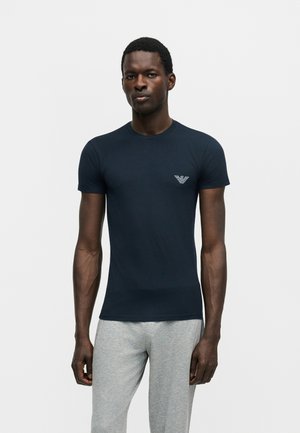 Muž nosi priliehavé tmavomodré tričko s malým bielym logom na hrudi a svetlosivé tepláky, stojaci na jednoduchom pozadí.