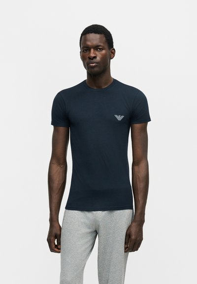 Άνδρας που φοράει εφαρμοστό μπλε σκούρο T-shirt με μικρό λευκό λογότυπο στο στήθος και ανοιχτό γκρι φόρμες, στέκεται μπροστά σε απλό φόντο.