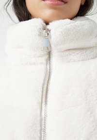 Chaqueta de forro polar blanca con cuello alto, cremallera plateada y textura suave. Presenta un material grueso y esponjoso para proporcionar calor.