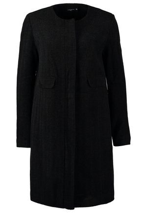 Femme marchant, portant un manteau noir, une chemise grise, un pantalon blanc, des bottines noires et tenant une pochette beige sur un fond uni.