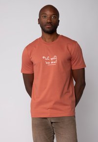 Homme avec une barbe courte portant un t-shirt de couleur rouille avec le texte "Ich zähl bis drei !" en blanc, se tenant devant un fond clair uni.