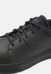 Zapatilla de cuero negro con acabado texturizado, punta redonda y cordones negros. Presenta un collar acolchado y un logotipo sutil en el costado. Suela de goma.