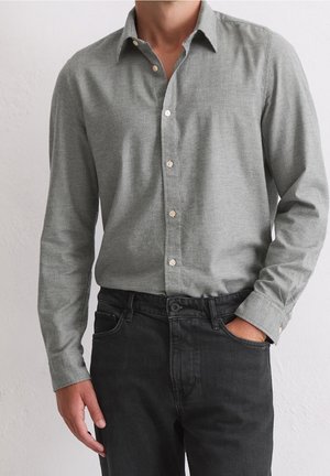Mann trägt ein hellgraues, langärmliges Hemd, das in schwarze Jeans gesteckt ist, mit einer Hand in der Tasche vor einem schlichten hellen Hintergrund.