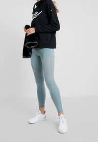 Felpa nera con logo bianco, abbinata a leggings verde menta con motivi testurizzati e design aderente. Scarpe da ginnastica bianche completano il look.