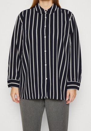 Persoon die een zwart overhemd met witte verticale strepen en een grijze broek draagt, staand tegen een effen witte achtergrond.